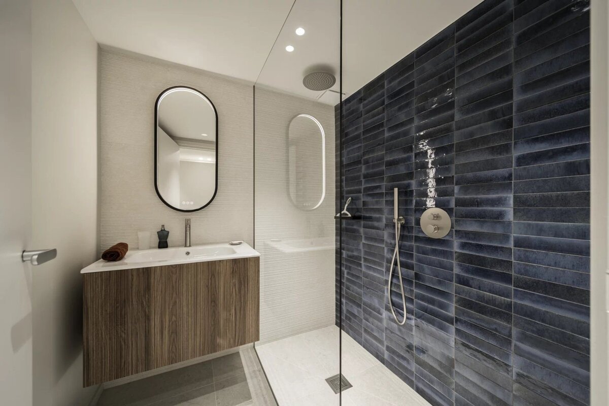 A sleek, modern bathroom features a dark blue tiled shower, glass partition, wooden vanity with a sink, and an illuminated oval mirror.