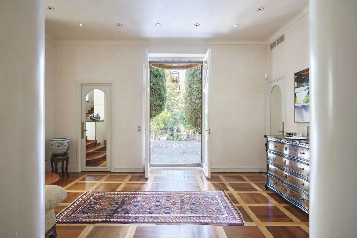 Elegant foyer with a patterned wood floor and rug. Double doors open to a beautiful garden. Features include an antique dresser and an arched doorway.