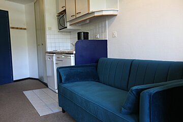A cozy studio apartment with a teal sofa facing a compact kitchen area, complete with white cabinets, appliances, and a tiled wall.