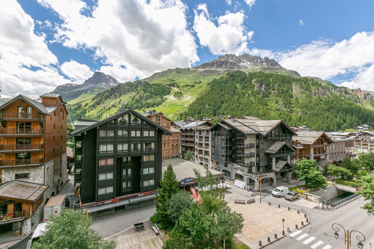 A picturesque alpine village with traditional wooden buildings and a town square, set against lush green mountains under a bright, cloudy sky.