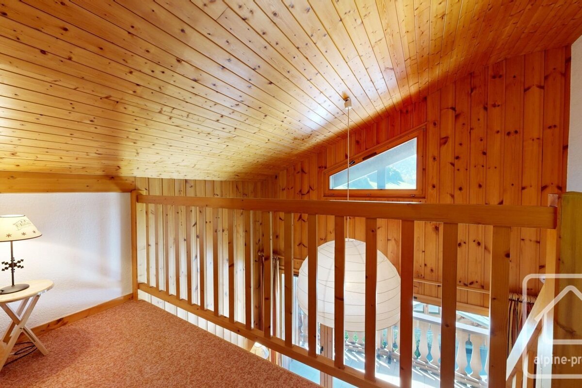 A cozy, wood-paneled loft features a wooden railing, triangular window, hanging paper lantern, and a small table with a lamp.