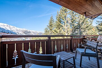 Cozy wooden chalet balcony with patio furniture overlooking snowy mountains and green pine trees under a clear blue sky.