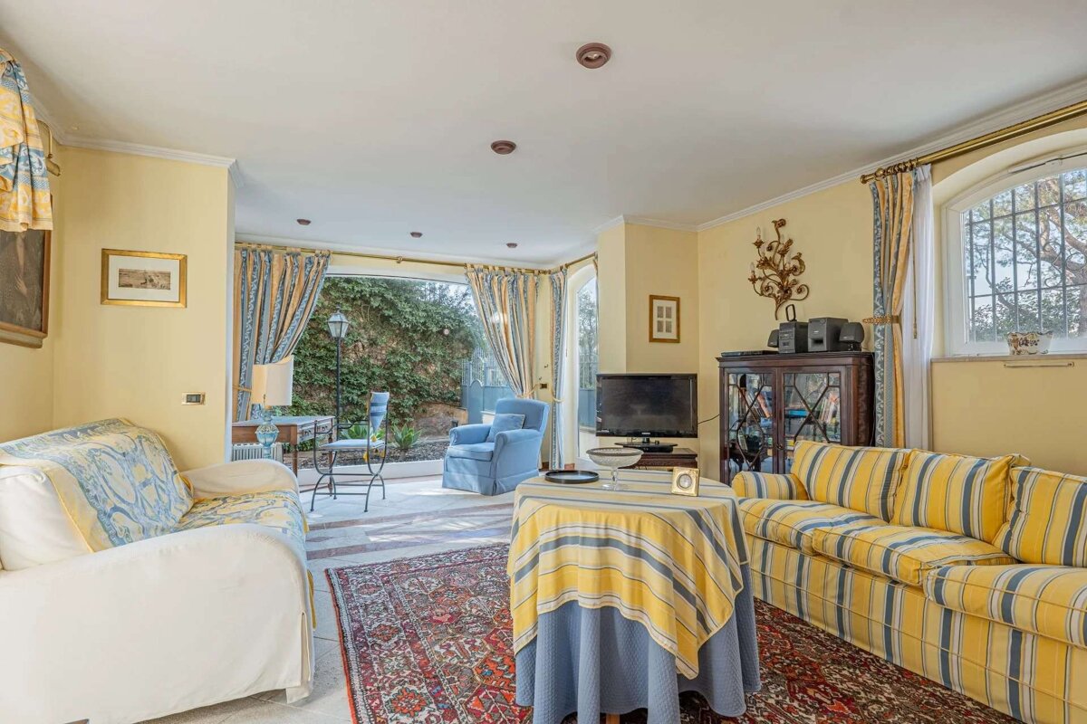 A bright, traditionally furnished living room with yellow walls, blue and yellow striped sofas, a patterned rug, and large windows looking out to a lush green garden.