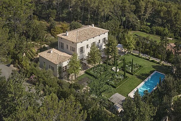 Aerial view of a large, light-colored villa with tiled roofs, nestled in a dense forest. Features include formal gardens, a terraced landscape, and a blue swimming pool.
