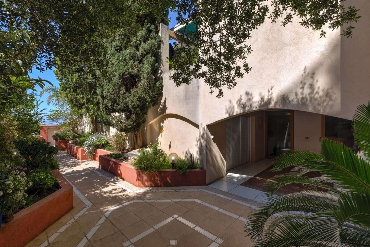 A sunny outdoor scene featuring a beige building with a curved entry, lush green trees, red planters, and a geometrically tiled patio dappled with shadows.