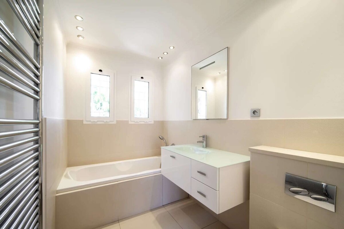 A minimalist bathroom with a bathtub, sleek floating vanity, large mirror, chrome towel warmer, and two small windows under recessed lights.