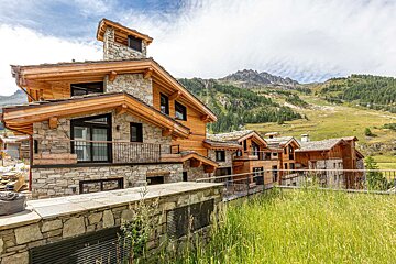 Rustic stone and timber chalets nestled on a grassy mountain slope under a cloudy sky, with distant peaks and pine trees.