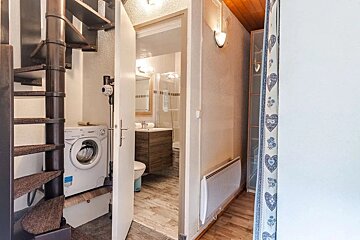 A compact utility space with a spiral staircase, washing machine, and vacuum. An open door reveals a modern bathroom, and a narrow hallway is visible right.