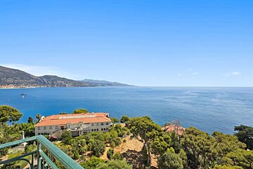 A picturesque Mediterranean villa amidst green trees, overlooking a vast blue sea with distant mountains under a clear sky, viewed from a balcony.
