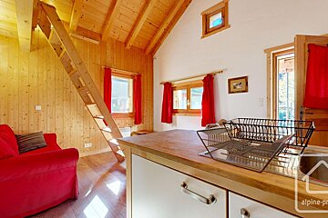 A sunlit, rustic cabin interior with wood walls/ceiling, red sofa, and a ladder. A kitchen counter with a dish rack is in the foreground, vibrant red curtains frame windows.