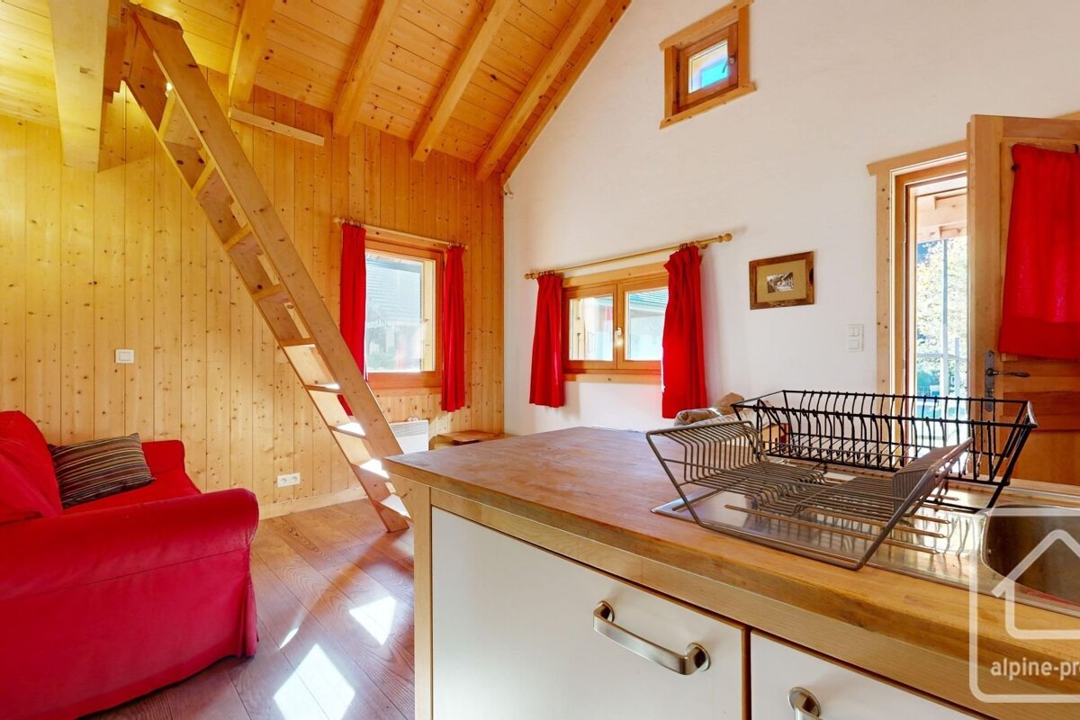 A cozy, sunlit room featuring wooden walls, a red couch, loft ladder, and a kitchen counter with a dish rack. Red curtains frame the windows.