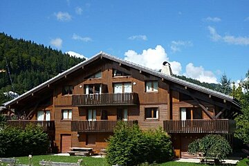 A large multi-story wooden alpine chalet with balconies, surrounded by green trees and hills under a clear blue sky.