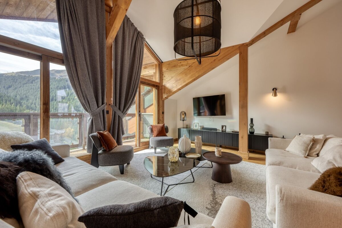 A modern rustic living room with large windows overlooking mountains. Features wooden beams, neutral sofas, and stylish decor.