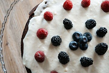 Close-up of a dark cake with white frosting, garnished with scattered red raspberries, black blackberries, and blue blueberries.