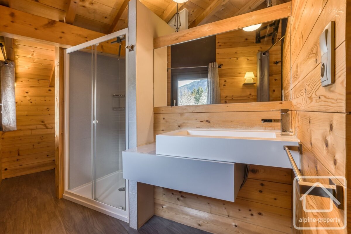 Cozy, modern bathroom in a wooden cabin featuring a white sink, glass shower, and a mirror reflecting a scenic mountain view.