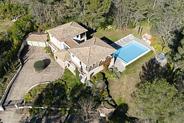 Aerial view of a sprawling villa with two buildings, a sparkling blue pool, large driveway, and multiple garages, surrounded by lush woodlands.