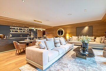 Luxurious modern living room with warm wood-paneled walls, plush sofas, a marble coffee table, and a dining area in the background.