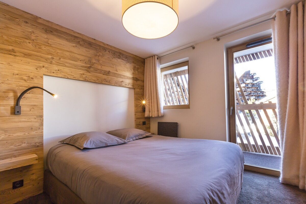 A cozy chalet bedroom with wood paneling, a large bed, and a glass door opening onto a balcony with snowy mountain views.