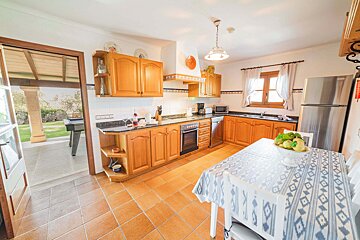 A bright kitchen with wooden cabinets, terracotta tiled floor, a dining table, and an open doorway leading to an outdoor patio.
