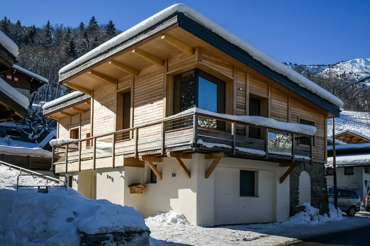 A modern wooden chalet in the snowy mountains under a bright blue sky, featuring a snow-covered roof and wooden balcony.