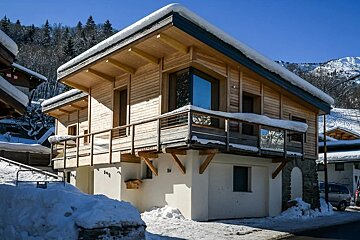 A modern two-story wooden chalet with snow-covered roofs in a mountainous region under a clear blue sky.