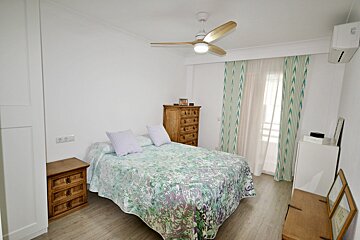 A bright bedroom with a bed featuring a green/purple patterned quilt, wooden nightstands/dresser, ceiling fan, and green patterned curtains.