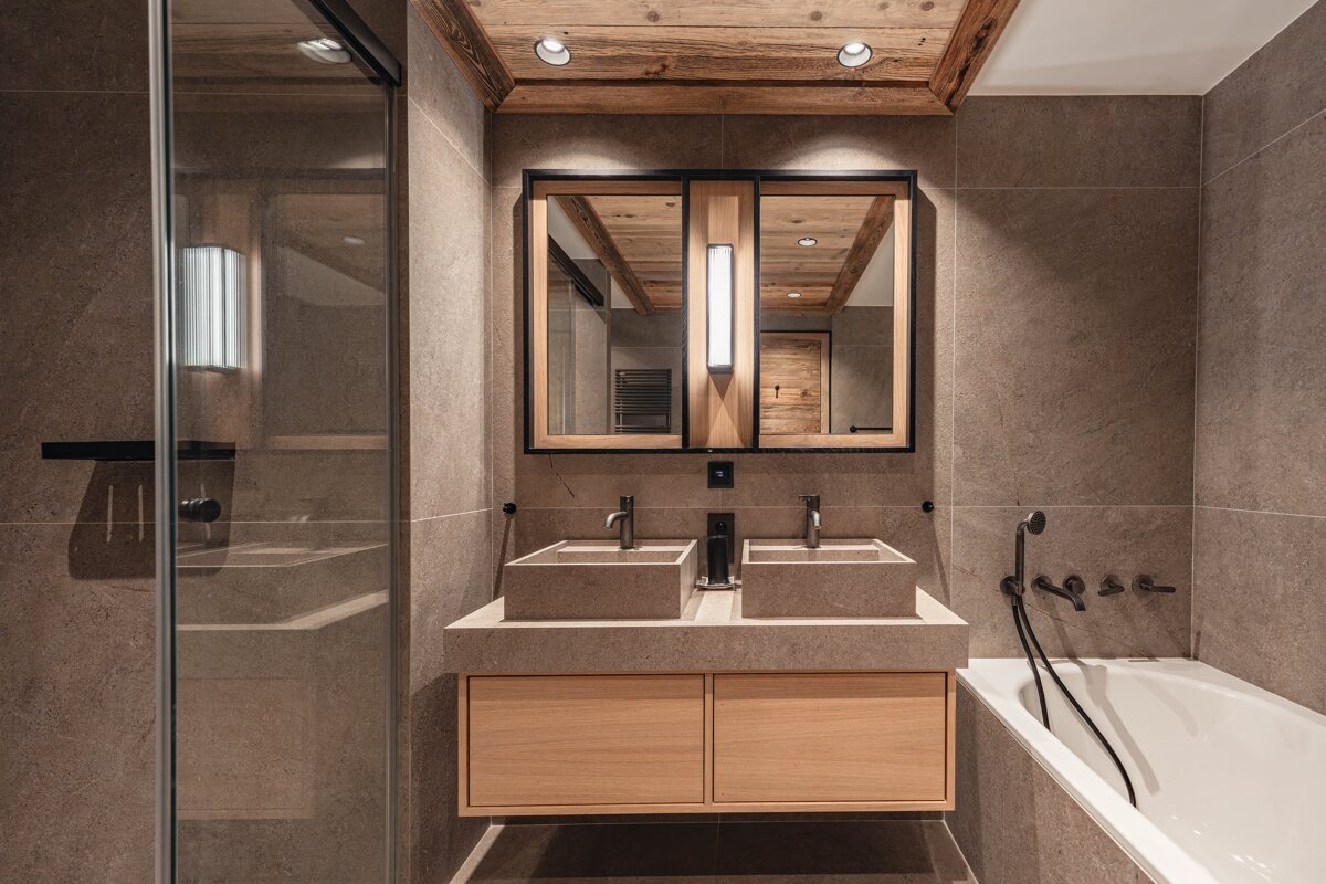 Modern rustic bathroom with double stone sinks on a floating wooden vanity, grey tiled walls, and a wooden ceiling.