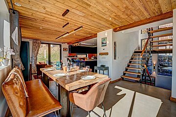 A modern, open-plan dining area with a rustic wooden ceiling, large timber table, and leather seating. A sleek linear light hangs above, with stairs and a kitchen visible.