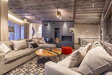 A modern, cozy living room features large grey sofas, a dark fireplace wall with wood storage, a rustic wooden ceiling, and a unique coffee table.