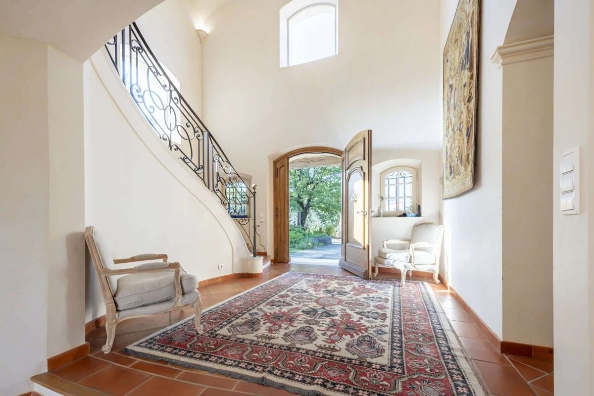A bright, grand entryway features a curved staircase with wrought iron railing, a large ornate rug on terracotta tiles, and open wooden doors leading outside.