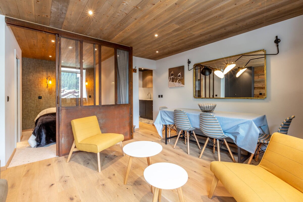 A modern, open-plan apartment featuring a bedroom behind a rustic glass partition, a dining area, and a glimpse of a kitchen. Warm wooden ceiling and floor.