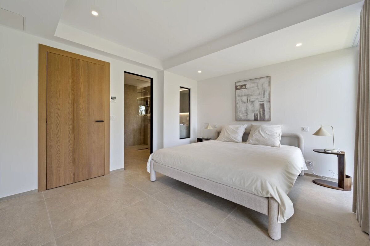 Minimalist modern bedroom with a large cream bed, tiled floor, wooden door, and an adjoining bathroom visible in the background.