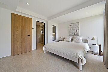 Minimalist modern bedroom with a large cream bed, tiled floor, wooden door, and an adjoining bathroom visible in the background.