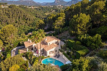 Aerial view of a luxurious villa with a swimming pool, surrounded by lush green trees and palm trees, set against a backdrop of mountains.