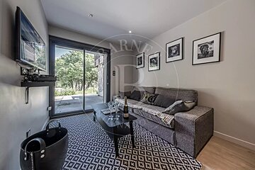 A modern living room with a grey sofa, black and white geometric rug, wall-mounted TV, and sliding glass doors revealing greenery outside.