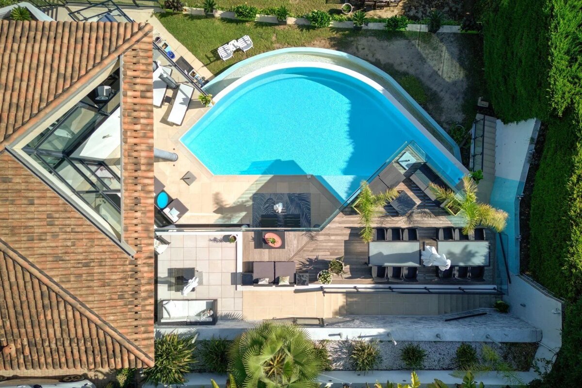 Aerial view of a luxurious property featuring a kidney-shaped swimming pool, multiple outdoor lounges on decks and patios, and lush landscaping.