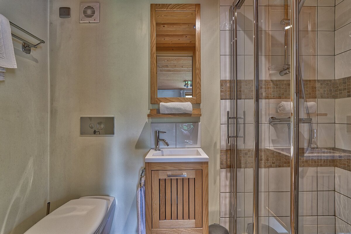 A compact, modern bathroom with a wood-framed mirror, vanity sink, partially visible toilet, and a glass shower with white and brown tiled walls.