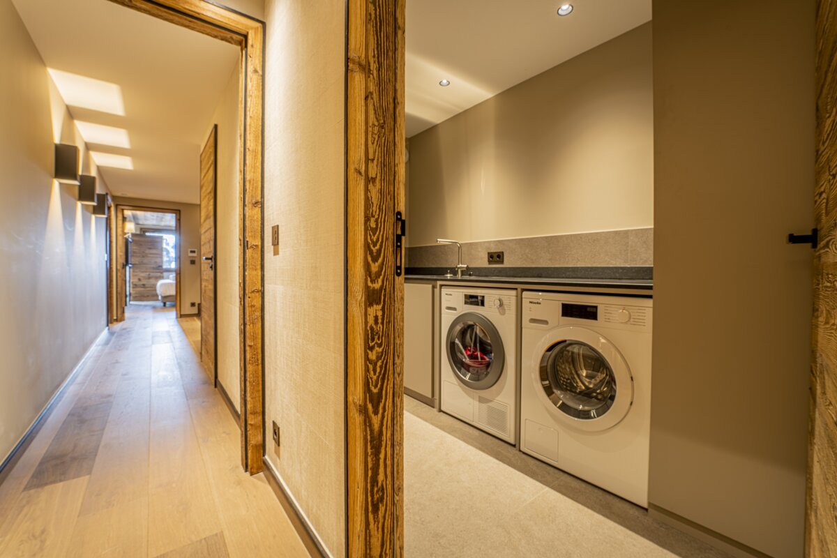 A modern hallway with light wood floors and rustic wood door frames, leading to a laundry room with two white washing machines and a sink.