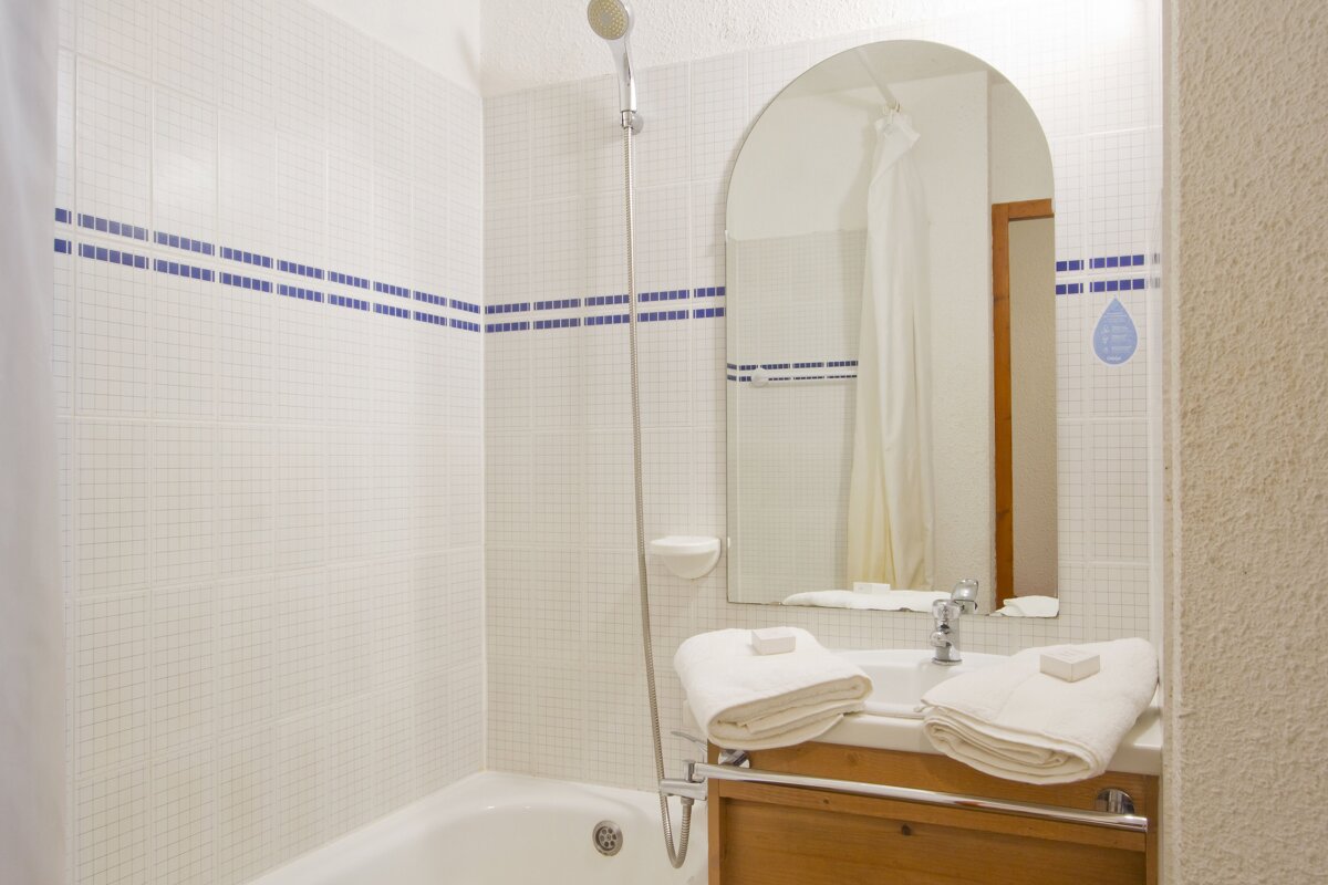 A clean, white-tiled bathroom featuring a bathtub with a shower, a sink with an arched mirror, and blue striped accents. Fresh towels are on the counter.