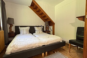 A cozy bedroom featuring two beds under a wooden staircase, with bedside lamps, white paneled walls, and a black chair.