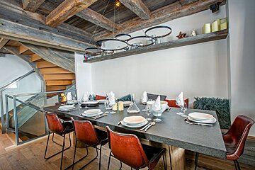 Rustic modern dining room with a long table set for guests, under exposed wooden beams and a circular chandelier. A wooden staircase is nearby.