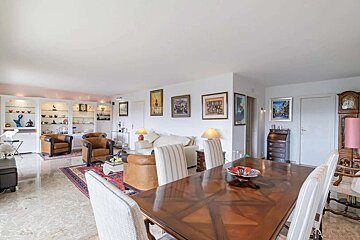 A bright, traditionally furnished living and dining room with white walls, marble floors, various artworks, display cabinets, multiple seating areas, and a large wooden dining table.