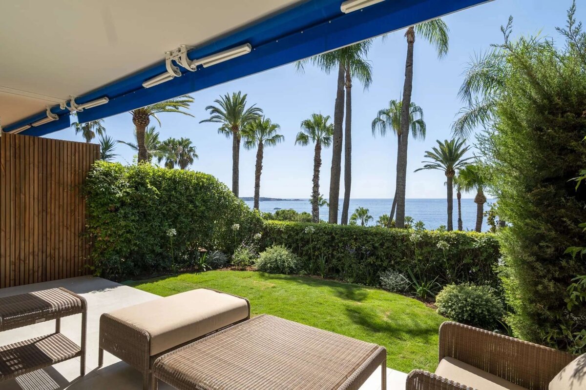 A sun-drenched patio with outdoor seating overlooks a vibrant green garden, tall palm trees, and the shimmering blue sea under a clear sky.