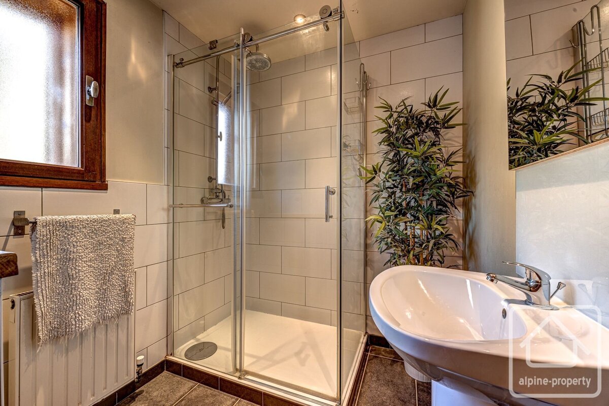 A bright bathroom with a glass shower, white basin, and a tall plant. Features white tiled walls, dark floor, and a wooden-framed window.