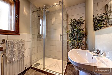 A bright bathroom with a glass shower, white basin, and a tall plant. Features white tiled walls, dark floor, and a wooden-framed window.
