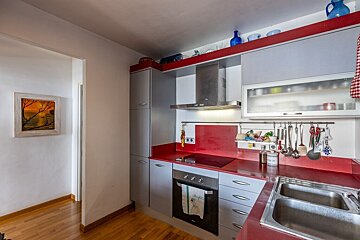 Modern kitchen featuring grey cabinets, vibrant red countertops, stainless steel appliances, and wood flooring leading to a hallway.