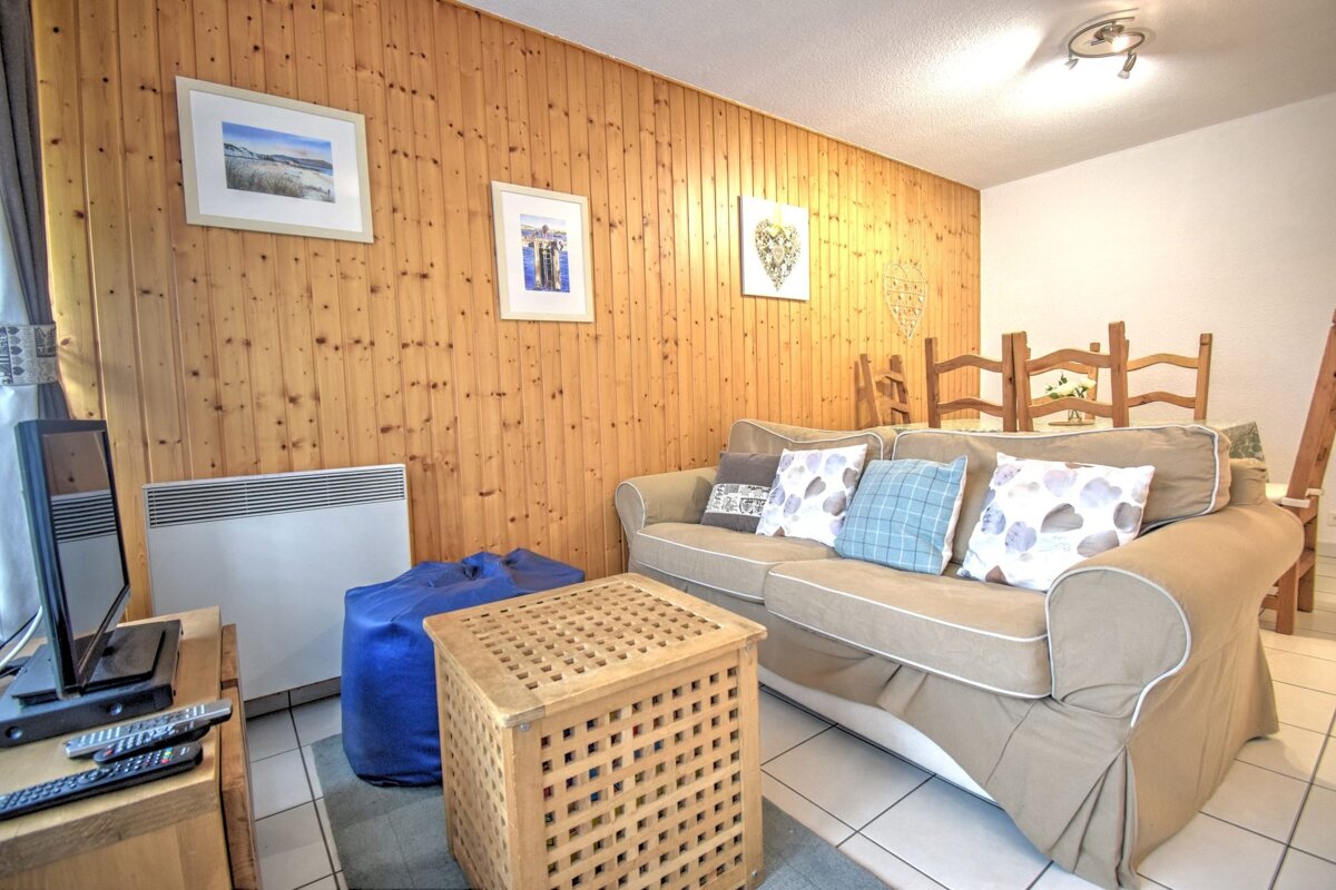 Cozy living room with a beige sofa, wooden paneled wall, lattice coffee table, blue beanbag, and a dining table in the background.