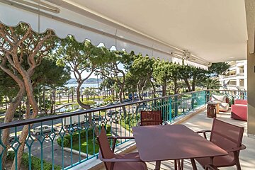 A sunny balcony with outdoor seating and an awning overlooks a tree-lined coastal street with traffic, a marina with boats, and distant hills.