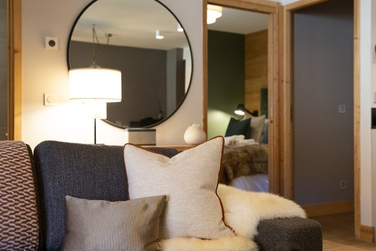 Cozy living room corner with a grey sofa, plush cushions, a large round mirror, and a view into a warm bedroom.