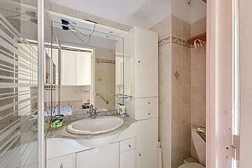 A bright, compact bathroom featuring a white vanity, sink, large mirror, tiled walls, white cabinets, and a frosted glass shower enclosure.